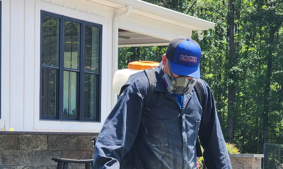 A Rozier Pest Control & Waterproofing technician applying a barrier spray to residential landscaping with a backpack sprayer.